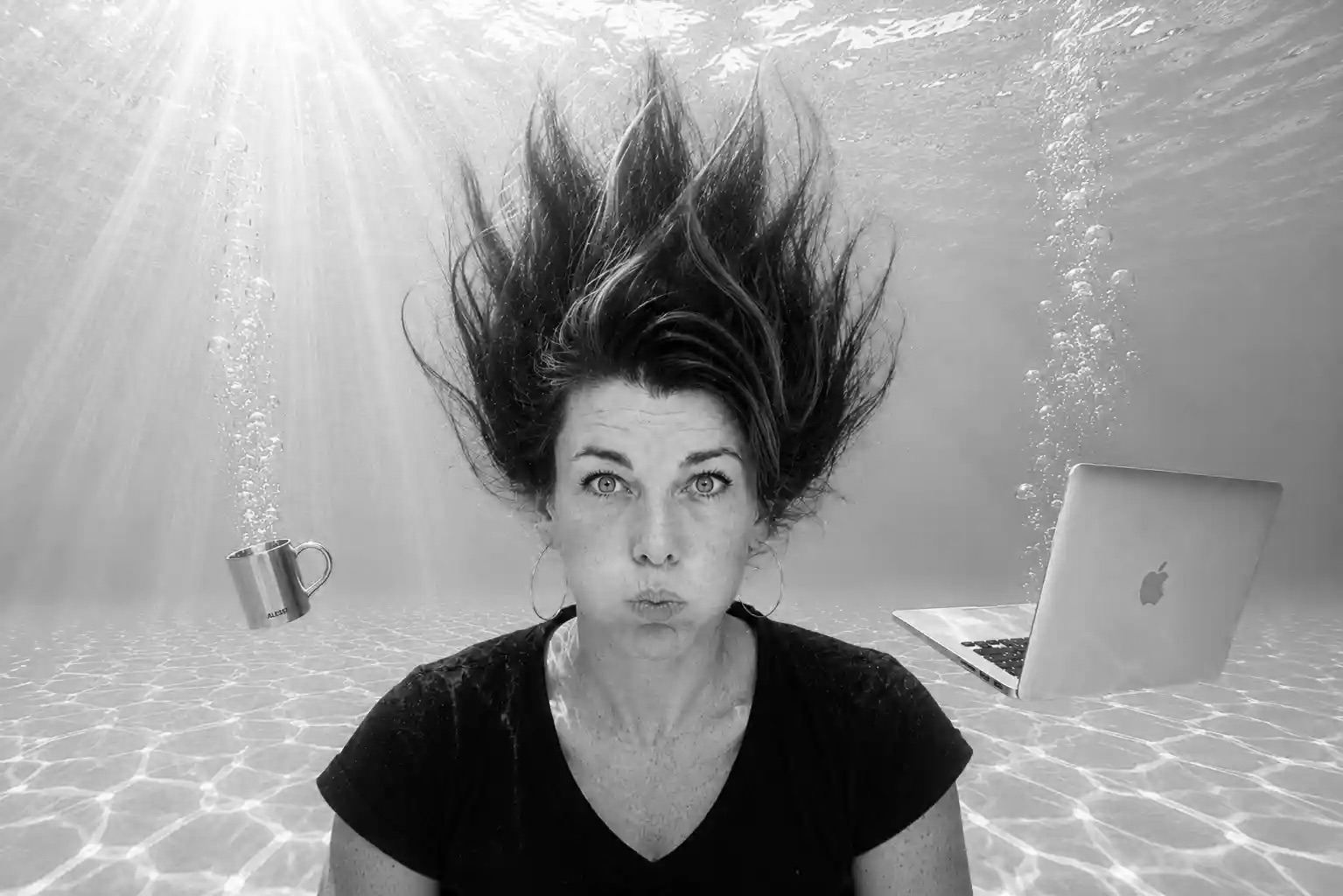 Joanne underwater, cheeks puffed, with a floating laptop and coffee cup